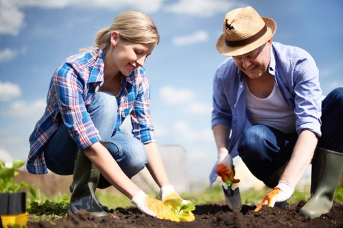 A young woman and an older woman are engaged in gardening in an outdoor garden space under a clear blue sky. The younger woman, dressed in a blue plaid shirt and jeans, is kneeling on the soil and planting or tending to a small leafy green plant, smiling as she works. The older woman, wearing a light blue jacket, white t-shirt, and a sun hat, is also kneeling and using a trowel to dig into the soil nearby. Both women are wearing gardening gloves and rubber boots, indicating they are actively involved in garden maintenance or planting. The garden features rich, dark soil in the foreground, with neatly prepared planting beds and healthy green plants. The background shows a landscaped yard or garden area, possibly including low hedges or shrubbery, with some planters or gardening tools nearby. The scene is bathed in natural sunlight, suggesting a pleasant day suitable for outdoor gardening activities, corresponding to residential gardening services offered by Garden Maintenance Islington in the N7 postal district or surrounding areas of London.