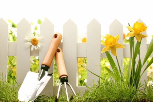 A close-up view of a garden with a white picket fence in the background, featuring bright yellow daffodils and white daisies growing along the fence line. The foreground shows lush, green grass with a slightly uneven texture, indicating recent mowing or trimming. Leaning against the fence are two gardening tools—a hand trowel with a wooden handle and a small garden fork—suggesting active garden maintenance. The garden appears to be part of a residential outdoor space, possibly a front or back garden in Islington, with natural sunlight illuminating the vibrant plant colours and casting soft shadows. The scene reflects a well-maintained, cheerful outdoor area suitable for gardening services offered by Garden Maintenance Islington, with a focus on lawn care, planting, and general garden upkeep.