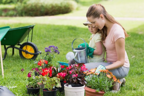 The image shows a woman and a young girl working together in a tidy, well-maintained garden in Islington, with a lush green lawn and various potted flowering plants such as geraniums and petunias arranged in the foreground. Behind them, a wheelbarrow with a yellow wheel is positioned on the grass, indicating gardening activities. The background features a neatly trimmed hedge and a soft, blurred lawn area, suggesting a peaceful outdoor space suitable for gardening and outdoor maintenance. Both individuals are engaged in planting or tending to the flowers, wearing casual clothing appropriate for gardening tasks. The scene reflects a bright, sunny day, with natural light illuminating the vibrant colours of the flowers and the green of the grass, demonstrating typical features of a well-kept, landscaped garden as serviced by companies like Garden Maintenance Islington.