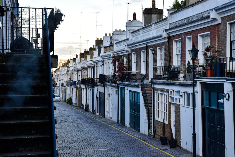 A row of traditional terraced houses with brick and painted facades, featuring small front gardens and balconies with metal railings. The buildings have white decorative cornices and sash windows, som