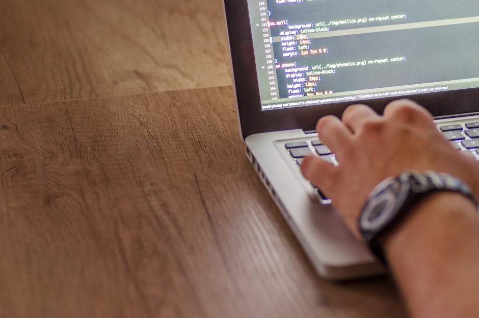 A close-up view of a person's hand operating a laptop on a wooden desk, with visible code or text on the screen. The individual is wearing a wristwatch with a black strap and silver face. The desk has
